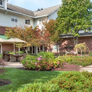 A landscaped garden area with a building in the background featuring trees, shrubs, and a patio with umbrellas.