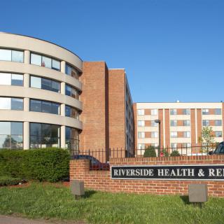 A healthcare facility with a sign reading 'Riverside Health & Rehab' in front of modern buildings under a clear blue sky.