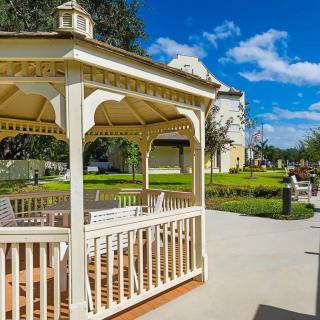 A sunny day in a park featuring a wooden gazebo, surrounded by trees and benches under a blue sky with clouds.