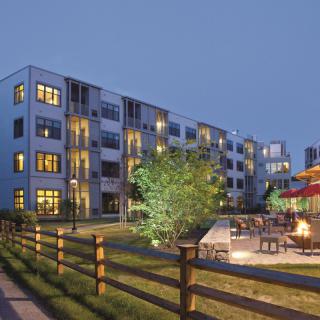 An illuminated apartment building with a fenced yard and outdoor seating area in the evening.