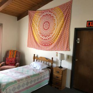 A cozy bedroom featuring a single bed, bedside table with a lamp, and a colorful tapestry hanging on the wall.