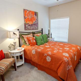 A cozy bedroom with vibrant orange bedding and decorative pillows, featuring a lampshade and artwork on the wall.