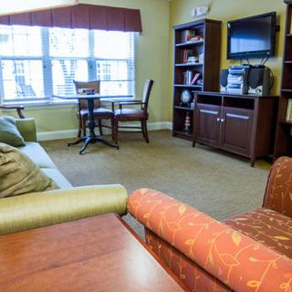 A cozy living room featuring a sofa, television, bookshelf, and a small dining table by the window.