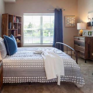 A cozy bedroom with a neatly made bed, bookshelves, and natural light coming through a large window.