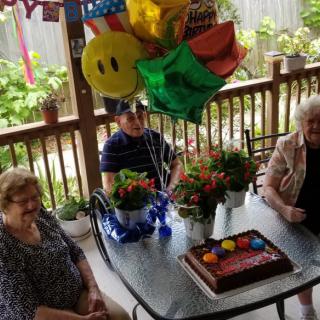 A group of people celebrating a birthday outdoors with balloons and a cake on the table.