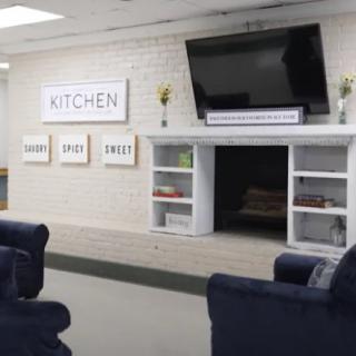 A cozy living area featuring a wall-mounted TV, comfortable seating, and decorative shelves in a kitchen-themed space.