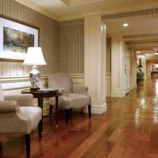 A well-lit hotel hallway with wooden flooring, featuring comfortable chairs, a side table with a lamp, and framed artwork on the wall.