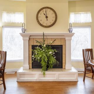 A cozy living room with a fireplace adorned with a clock and decorative plants, flanked by large windows and wooden chairs.
