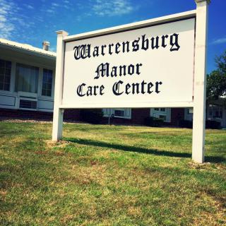 A sign for Warrensburg Manor Care Center stands on a grassy lawn under a clear blue sky.
