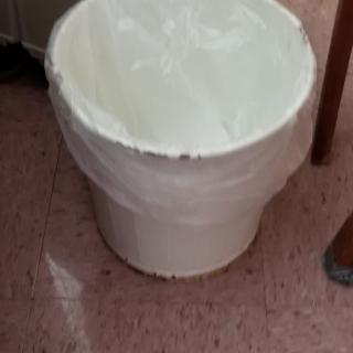 A trash can with a plastic bag liner placed on a tiled floor in an indoor setting.