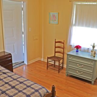 A cozy bedroom with wooden flooring, featuring a bed, chair, and dresser near a sunlit window.
