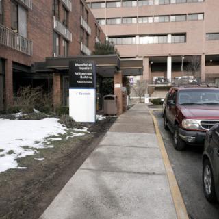A view of a hospital entrance with a sidewalk and parked cars, showing some snow on the ground.