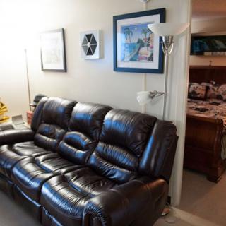 A cozy living room with a leather sofa, wall art, and a floor lamp, leading into a bedroom.