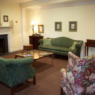 A cozy living room with traditional furniture and decor, featuring a green sofa, floral armchair, and a coffee table set on a carpet.