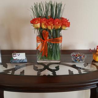 A decorative arrangement of orange roses in a vase with a matching ribbon, displayed on a glass-top table.