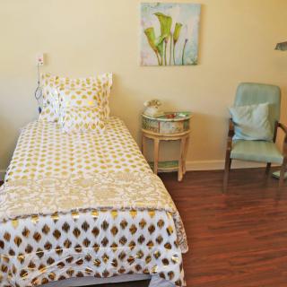 A cozy bedroom with a single bed, patterned bedding, a nightstand, and a comfortable chair on a wooden floor.