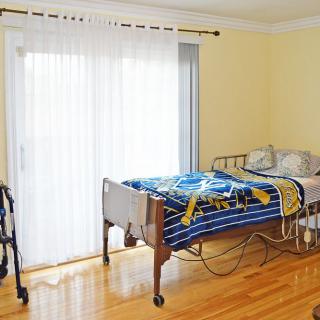 A bedroom featuring a hospital bed, walker, and large window with blinds in a bright space with hardwood floors.