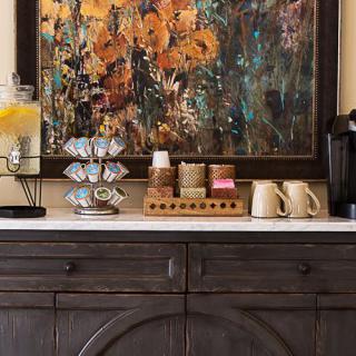 A cozy coffee station featuring a coffee maker, assorted cups, and a lemon water dispenser, all set against a colorful abstract painting.