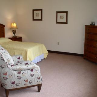 A cozy bedroom with a bed, patterned chair, wooden dresser, and soft lighting.