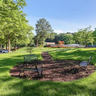 A scenic view of a park with benches arranged around a pathway, surrounded by lush greenery and trees on a sunny day.