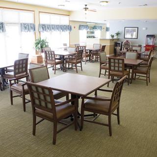 A spacious dining area with multiple tables and chairs arranged on a carpeted floor, well-lit by natural light.
