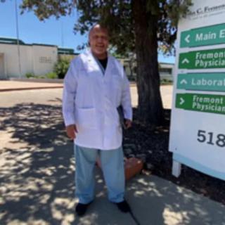 A person wearing a lab coat stands outside near a sign and tree.