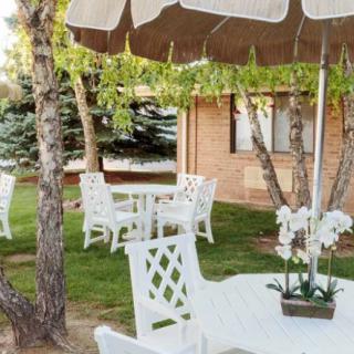 A peaceful garden area featuring white patio furniture with umbrellas set amidst greenery and trees, adjacent to a brick building.