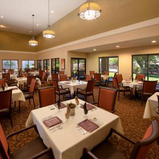 A spacious and elegantly decorated dining room with multiple tables set for guests, featuring warm lighting and large windows overlooking the garden.