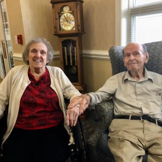 An elderly couple sitting together in a cozy living room, holding hands and smiling.