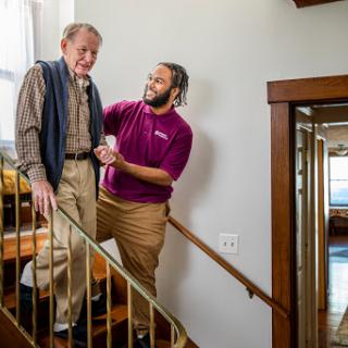 A caregiver provides support to an elderly man as they navigate the stairs together.