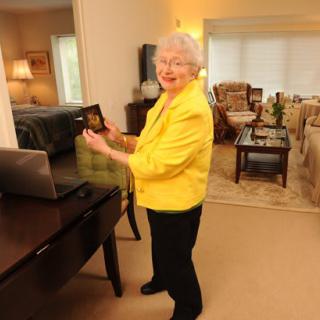 An elderly woman smiling in her living room, wearing a yellow jacket and standing near a laptop.