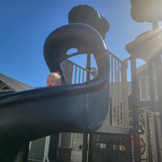 A child peeks out from the top of a playground slide under the clear blue sky.