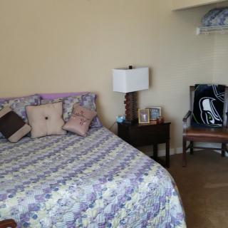 A cozy bedroom with a patterned bedspread, pillows, nightstand with a lamp, and a chair in the corner.