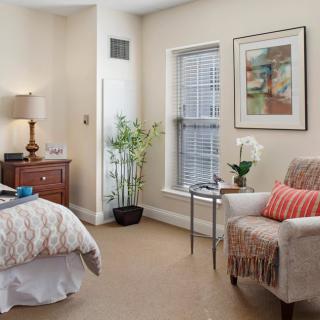 A cozy bedroom with elegant decor, featuring a bed, armchair, side table, and potted plants.