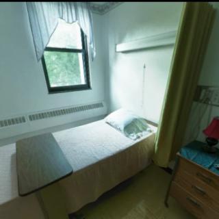 A small, simple room with a single bed next to a window, accompanied by a bedside table and dresser.