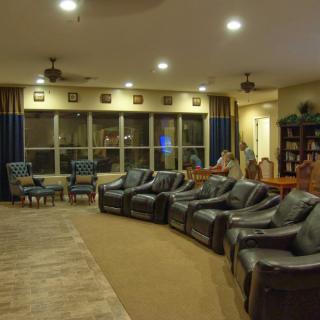 A spacious living room featuring a row of recliner chairs, large windows with curtains, and cozy ambient lighting.