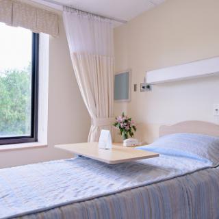 A serene and well-lit hospital patient room featuring a bed with a light blue blanket, a small table, and a window adorned with curtains, complemented by a vase of fresh flowers.