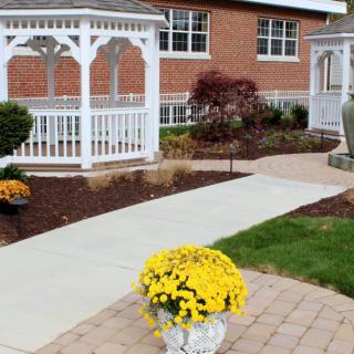 A well-maintained garden featuring a white gazebo and colorful potted flowers along a walkway next to a brick building.
