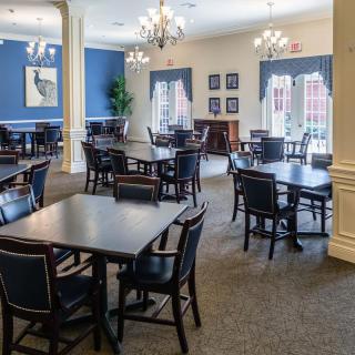 A spacious dining area with multiple tables and chairs, featuring elegant chandeliers and a carpeted floor, complemented by blue accent walls and large windows.