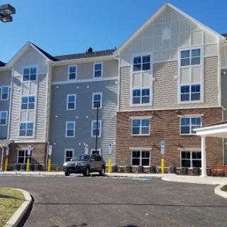 A modern hotel building featuring a combination of siding and brick exterior, with a spacious parking area under clear blue skies.
