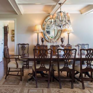 A traditional dining room featuring a crystal chandelier, a long wooden table with matching chairs, and an ornate mirror on the wall.