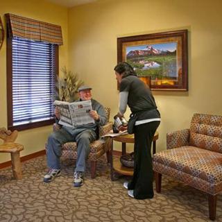 A person reads a newspaper in a cozy waiting room while another individual assists them.