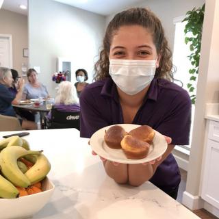 A woman wearing a mask holds a plate of muffins in a kitchen setting.