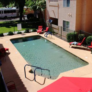 An outdoor swimming pool at an apartment complex on a sunny day, surrounded by lounge chairs and a safety fence.