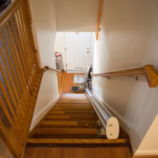 A view down a wooden staircase with railings on both sides, leading to a well-lit hallway.