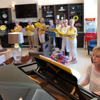 A group of women singing in a choir while one plays the piano indoors.