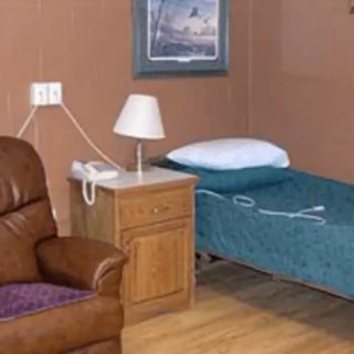 A simple hospital room featuring a bed, nightstand with a lamp, and a comfortable chair.