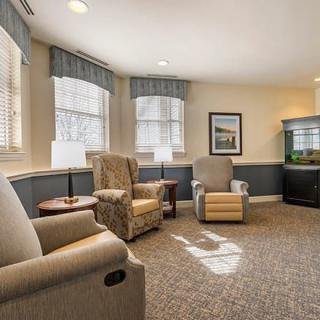 A cozy living room with armchairs and a carpeted floor, well-lit by natural light from the windows.