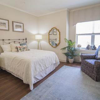 A cozy bedroom with neutral tones, featuring a neatly made bed, a plush armchair, a potted plant, and natural light streaming through the window.