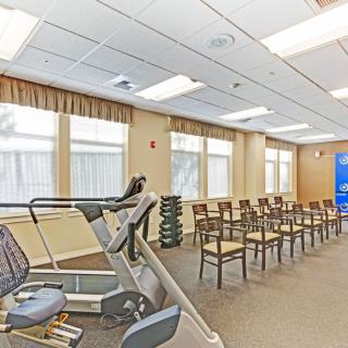 A multipurpose room with gym equipment and rows of chairs, illuminated by natural and ceiling lights.